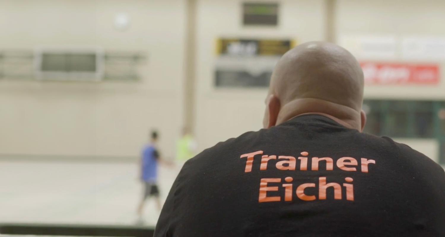 Eine Person mit Glatze ist von hinten zu sehen und trägt ein schwarzes T-Shirt mit der orangefarbenen Aufschrift ‚Trainer Eichi‘. Im Hintergrund befindet sich eine unscharfe Sporthalle, in der zwei Personen auf dem Spielfeld stehen. Eine Person mit Glatze ist von hinten zu sehen und trägt ein schwarzes T-Shirt mit der orangefarbenen Aufschrift ‚Trainer Eichi‘. Im Hintergrund befindet sich eine unscharfe Sporthalle, in der zwei Personen auf dem Spielfeld stehen.