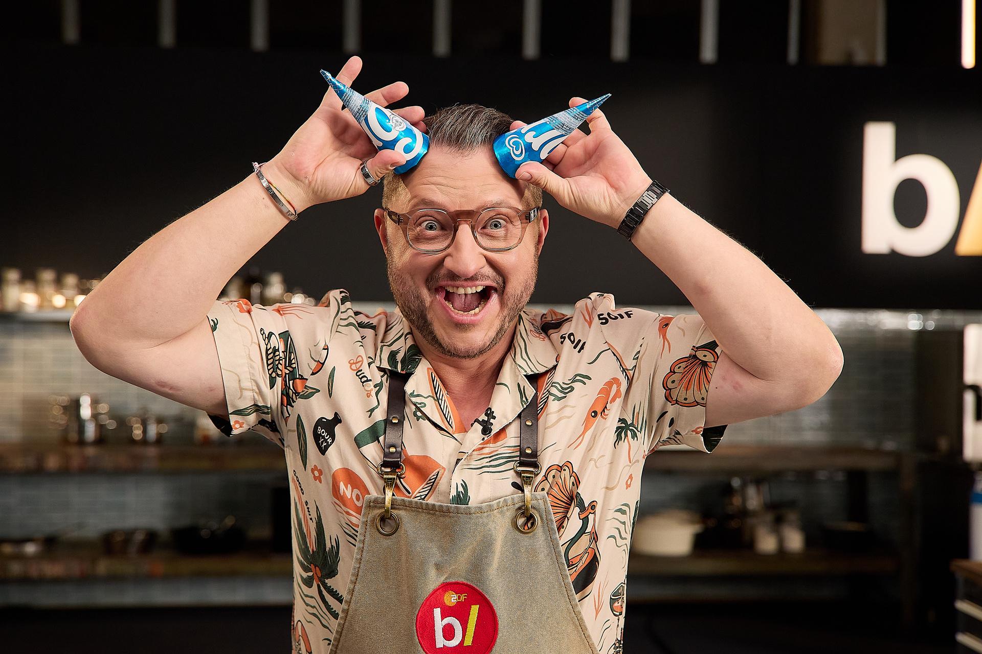 Sebastian Lege steht in einer Küche und trägt eine Schürze mit dem besseresseer-Logo und hält zwei blaue Eistüten als Hörner über der Stirn. Im Hintergrund sind Regale und Küchengeräte zu sehen. Sebastian Lege steht in einer Küche und trägt eine Schürze mit dem besseresseer-Logo und hält zwei blaue Eistüten als Hörner über der Stirn. Im Hintergrund sind Regale und Küchengeräte zu sehen.